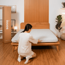 Woman placing mattress on Sonno wooden bed frame, emphasizing easy assembly and comfort in modern bedroom setup.